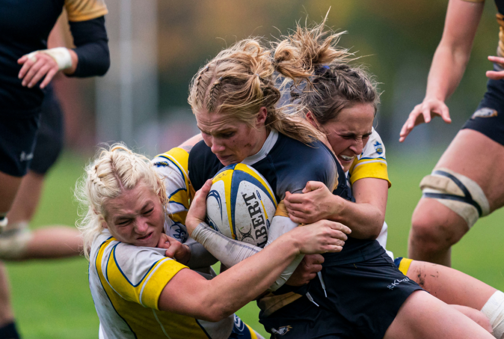 A female rugby player holding a Rugby ball is tackled by two opposing players
