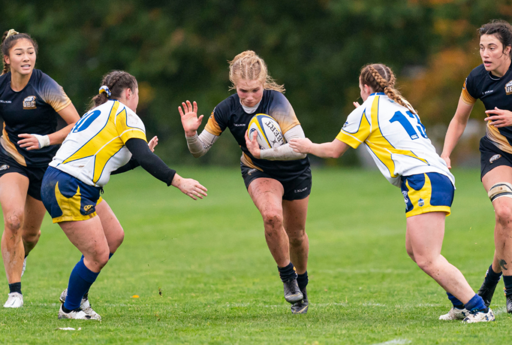 A female rugby player runs with the ball through two opposition players