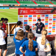 A group of children listen to someone giving a talk in front of a media advertising board
