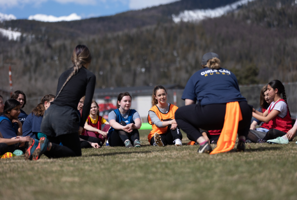 A group of girls sit on a field as a coach gives instructions. There is a mountain in the background