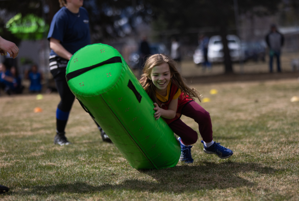 A close up of a girl running on grass and tackling a green tackle bag