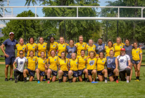 A group of female rugby players in gold kit pose for a team photo under a set of Rugby posts