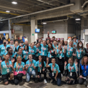 A group of volunteers dressed in matching teal shirts pose and wave at the camera