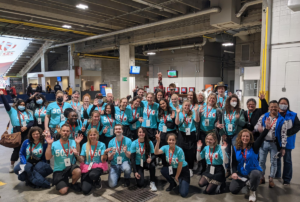 A group of volunteers dressed in matching teal shirts pose and wave at the camera