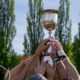 A close up of a team lifting a trophy with a blurred background of trees