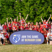 A group of girls pose for a Rugby team photo behind a blue banner that reads Join The Game at BCRugby.com