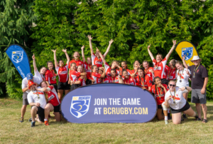 A group of girls pose for a Rugby team photo behind a blue banner that reads Join The Game at BCRugby.com