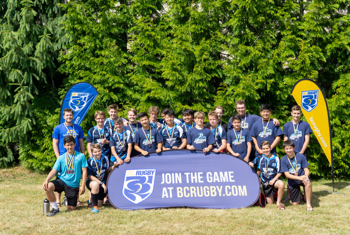 A group of boys pose for a Rugby team photo behind a blue banner that reads Join The Game at BCRugby.com