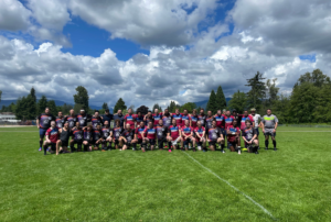 A group of Rugby players pose for a team photo in the middle of a green pitch. There are trees and mountains in the background.