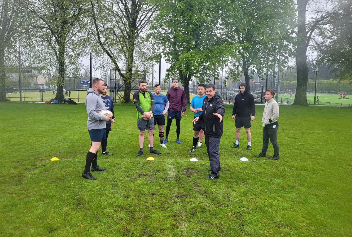 A Rugby coach demonstrates a drill to a group of Rugby players on a field