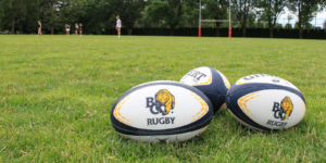 An image of three BC Rugby branded Rugby balls on a field with blurred players in the background