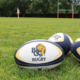 An image of three BC Rugby branded Rugby balls on a field with blurred players in the background