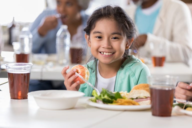 A young child sits in the forefront of a photo eating an apple and salad