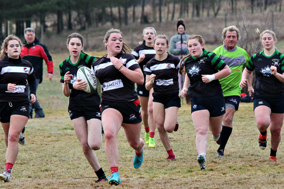 A female player runs with a rugby ball with a chasing pack of players behind